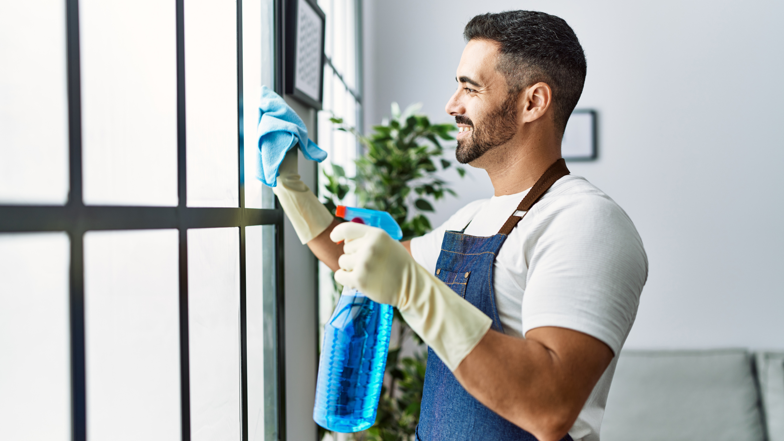 A smiling man wearing a white t-shirt, apron, and rubber gloves is cleaning a window with a blue cloth and a spray bottle in a bright indoor setting.